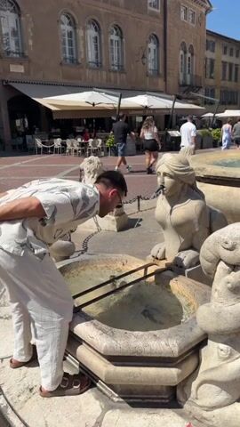 A man wearing sunglasses, a patterned short-sleeved shirt, and white pants is bending over a stone fountain. The fountain has decorative carvings of mythical creatures. Water is flowing from a spout into the basin of the fountain. The man appears to be drinking water from the fountain. In the background, there is a paved square with tables and chairs outside a building with awnings. Several other people are visible in the background, some walking and some seated at tables. The scene appears to be an outdoor public space in a European town or city, possibly during the daytime given the lighting.
