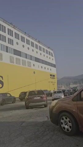 A fleet of cars is parked on a paved surface next to a massive yellow and white ship. The ship has multiple decks with numerous windows. The sky is clear and blue. The cars are of various makes and models, and they appear to be waiting to board the ship or disembark. Some of the cars are facing forward, while others are facing backward. The video is shot from a low angle, making the ship appear even larger. The overall impression is one of a busy port with a lot of activity.