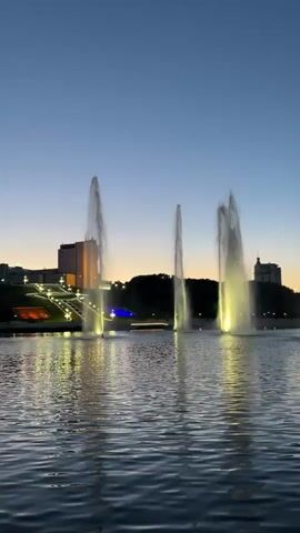 The video captures a wide shot of a body of water at dusk, featuring several large illuminated fountains. The fountains spray tall jets of water, which are lit from below with a warm yellow-white light, reflecting on the rippling surface of the water. In the background, a cityscape is visible, with buildings and illuminated structures on both sides of the water. The sky transitions from a deep blue overhead to a lighter orange hue at the horizon. No people are visible.