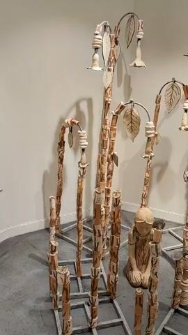 A video shows an art installation featuring tall, slender, light brown and white vertical sculptures arranged in a grid-like pattern on the floor. In the center of the grid, a light brown sculpture of a kneeling person with their head bowed is visible. Some of the taller sculptures have leafy and floral elements at their tops, created with the same light brown and white materials. The sculptures are set against a plain, light-colored wall.