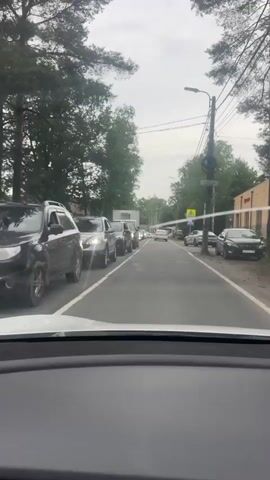 The video shows a view from inside a car looking forward through the windshield. The car is in traffic on a two-lane road lined with trees. Several cars are visible ahead, stopped or moving slowly. The sky is overcast. A traffic light pole with wires is visible on the right side of the road. A yellow pedestrian crossing sign is also visible on the right.