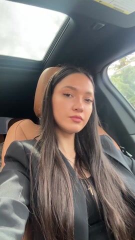 A young woman sits in the driver’s seat of a car with brown leather seats and a sunroof, wearing a black blazer over a black top, a gold chain necklace, and a headband. She has long, straight, dark brown hair and is wearing lipstick. The camera is recording a close-up view, alternating between upright and sideways angles. The woman makes a kissing face towards the camera. Outside the car window, trees can be seen.