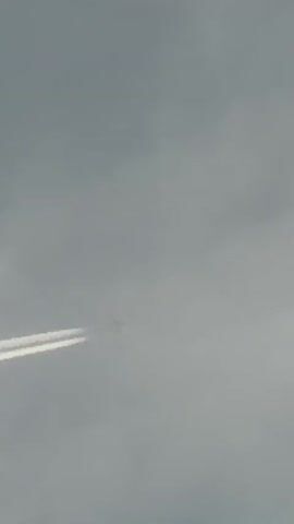 The video shows a plane flying overhead, leaving a contrail across the sky. The camera pans up from a gray, cloudy sky to reveal a house with a light brick exterior, a blue trim and a balcony. A tree is seen in the corner of the frame.