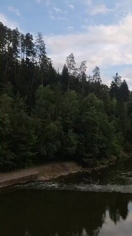 The video shows a view of a river flowing through a forested area. The trees are dense and green, covering the slopes of hills that rise on either side of the river. The sky above is partly cloudy with patches of blue visible. In the foreground, the dark water of the river reflects the trees and the sky. There is a small section of concrete embankment with steps visible on the left side of the river. Further down the river on the right bank, two concrete structures are partially submerged in the water, possibly remnants of a bridge or dam. The overall scene is peaceful and natural, with the sound of flowing water accompanying the visuals.