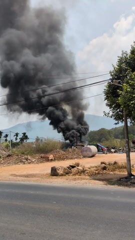 The video shows a large plume of black smoke rising into the sky. The source of the smoke appears to be on the ground, amidst a collection of rocks, equipment, and some vegetation. A large white cylindrical tank is visible on the ground nearby. The background includes a dirt road, power lines, and a mountain range in the distance with some palm trees.  A motorcyclist rides past on the road in front of the smoke.