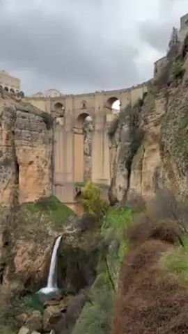 The video shows a view of the Puente Nuevo bridge in Ronda, Spain. The bridge spans a deep gorge, and a waterfall flows into the pool at the bottom. The cliffs of the gorge are covered in greenery. The sky is overcast.