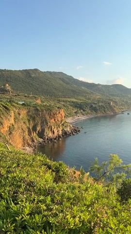 A slow, panning video showcases a scenic coastal landscape. The foreground features lush green vegetation with hints of cacti. The middle ground reveals a calm, blue sea meeting a rugged coastline with a small, secluded beach. In the distance, rolling green hills and mountains stretch along the horizon, bathed in the warm glow of a setting sun. A single sailboat is visible on the water. The sky is a clear, pale blue, transitioning to a soft orange hue near the horizon.