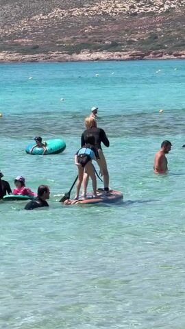 A group of people are enjoying a sunny day at the beach. In the foreground, a woman on a paddleboard is holding a paddle and appears to be teaching a young person standing behind her. Another person is partially submerged in the water next to the paddleboard. In the midground, a child is in a green inflatable tube, and another child is in a pink inflatable device. In the background, a man is swimming in the turquoise water. The sea is clear and calm, with a hilly, rocky shoreline in the distance. Yellow buoys are visible in the water.