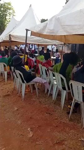The video shows a crowd of people sitting in chairs under white tents at an outdoor event. The ground is red dirt. Most people are seated, facing forward, watching something happening in front of them. The attendees are wearing colorful clothing. White plastic chairs are arranged in rows. There is lively music playing in the background.
