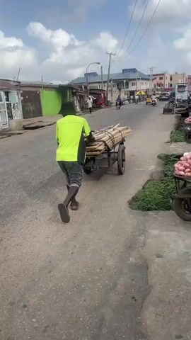 The video shows a street scene in what appears to be a West African country. A man wearing a bright green shirt and shorts is pulling a cart loaded with long, thin bundles of wood down the street. A white car is parked on the left side of the road. There are several motorcycles and other cars driving on the street in the distance. On the right side of the street, there are two carts filled with what appears to be onions or potatoes. There are a few small shops or stalls along the side of the road.  Later, a blue-green Toyota Corolla drives past. Two men are walking on the side of the road. The sky is cloudy.