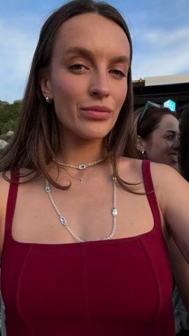 A woman with long brown hair, wearing a red dress and a silver necklace, smiles and winks at the camera. She is at an outdoor event, possibly a concert or festival, with other people in the background. The atmosphere appears to be lively and casual. There are drinks and snacks visible on a table in front of her. The woman is the primary focus of the video.