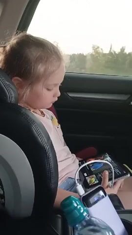 A young girl with blonde hair is sitting in the back seat of a car, looking down at a smartphone. She is wearing a light pink t-shirt with a yellow fruit design on it and a necklace. Her hair is pulled back in a bun, and sunlight is shining through the car window, illuminating her hair. A black leather car seat is visible in the foreground, and a portable charger is connected to the smartphone, providing power. A water bottle with a green cap is also visible in the foreground. The background shows a blurry view of trees and the sky, suggesting the car is in motion.