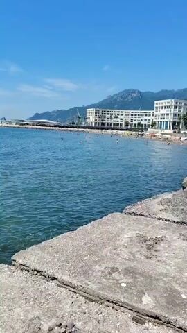 The video captures a view of a coastline with a blue sky, calm blue water, and mountains in the distance. In the foreground, there are concrete structures. Along the shore, a white building with multiple floors is visible, with people on the beach in front of it. Further out in the water, several individuals can be seen swimming or relaxing on floats. A harbor with cranes and sailboats is also present on the left side of the frame. The overall scene depicts a sunny day at a coastal resort or town.