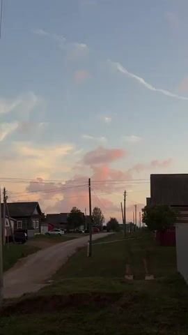 The video shows a rural village at dusk. The sky is filled with pastel-colored clouds, pink and orange hues mixing with the soft blue. A dirt road winds through the village, flanked by houses with simple architecture and tiled roofs. Wooden utility poles with power lines crisscross the scene, casting shadows as the light fades. In the foreground, grass and dirt cover the ground, and a portion of a vehicle is visible. A yield sign stands at an intersection, indicating traffic rules within this quiet setting. The overall atmosphere is peaceful and serene, capturing the beauty of a rural sunset.