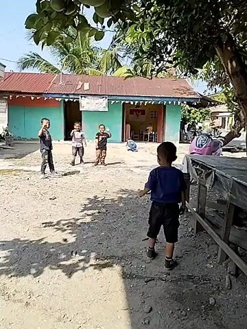 The video shows a group of four young children playing in an outdoor area in front of a building. The building appears to be a single-story structure with a red corrugated metal roof and teal-colored walls. There is a doorway in the center of the building, with some people visible inside. The ground is covered in dirt and gravel, with scattered shadows from trees overhead. One child, wearing a blue shirt and black shorts, stands facing away from the camera, observing the other three children. The other three children, also young boys, are playing further away, closer to the building. One of them is running, while the other two appear to be interacting. There are trees and vegetation around the area, suggesting a rural or semi-rural setting. A table with a dark tarp is visible in the foreground on the right side of the frame. A motorcycle is parked near the building on the right. The lighting suggests it is daytime, with bright sunlight.