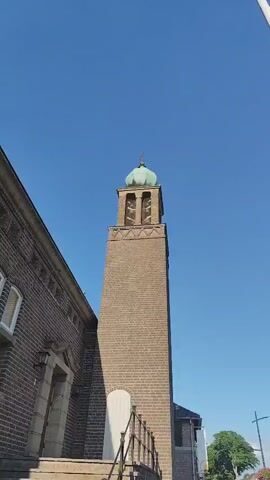 The video pans upwards along a tall brick bell tower on a clear, sunny day. The tower is topped with a distinctive teal-colored, rounded dome that has a cross on its very peak. Below the dome, the tower opens into a belfry where multiple bells are visible, protected by mesh netting. A decorative brick band with a zigzag pattern runs horizontally around the tower just below the belfry. The overall scene suggests a tranquil, possibly late morning or early afternoon, given the bright sunlight and clear blue sky. The architecture points to a European setting, consistent with the provided location context of Schaijk, Netherlands. There is no visible text in the video.