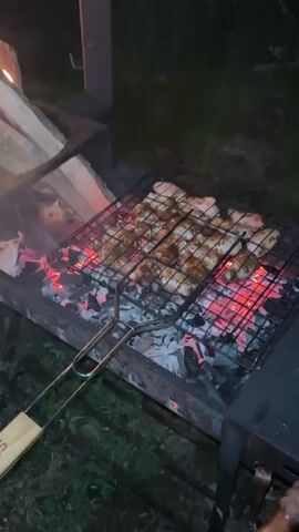 A full shot shows a person cooking meat on a grill over hot coals, with a fire burning in a separate section of the grill. The meat, likely chicken, is arranged on two rectangular grates positioned over glowing red coals. The flames from the fire cast an orange glow, illuminating the logs within the separate section. The overall scene is captured at night, with dark surroundings. The person's left hand is visible at the bottom of the frame, holding the handle of one of the grilling grates, which has a brand name "Stella" written on it. The camera slowly pans up and down during the video.