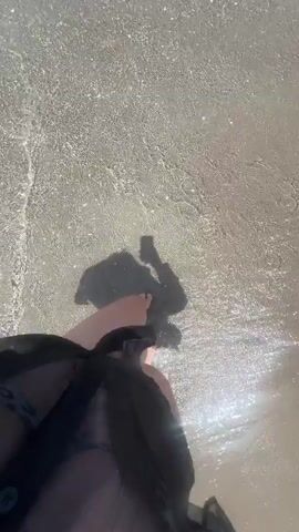 A low-angle shot, likely from a mobile phone, shows a person standing on a sandy beach as gentle waves wash around their feet. The person is wearing a dark, possibly sheer, garment that billows around their legs. A glimpse of swimwear is visible underneath. The camera casts a shadow on the sand. The water is clear and reflects sunlight, creating a shimmering effect.