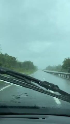The video shows a view from inside a car driving on a highway in the rain. The wipers are active, clearing water from the windshield. The road is wet and shiny, reflecting the overcast sky. Trees and grass line the side of the road. There is a guardrail on the right side of the highway. Visibility is reduced due to the rain.