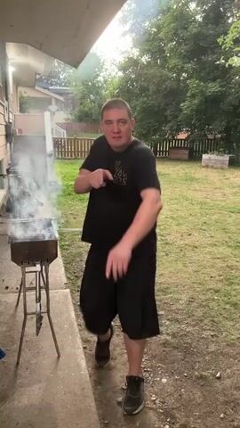 A man is grilling food on a charcoal grill in a backyard. He is wearing a black t-shirt and black shorts. He is holding a grilling tool with food on it and he is waving at the camera. There is smoke coming from the grill and the man is smiling. The backyard is grassy and there are trees and a fence in the background.