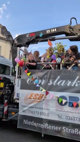 The video shows a group of people on the back of a truck with a crane. The truck is decorated with banners and balloons. The people appear to be celebrating or participating in a parade. One person is holding a phone and taking a picture. Another person is holding a drink. The banner on the side of the truck says "Ein starkes Team" which translates to "A strong team". There are also company names and phone numbers listed on the banner. The sky is blue and there are some buildings in the background. The overall mood seems festive.