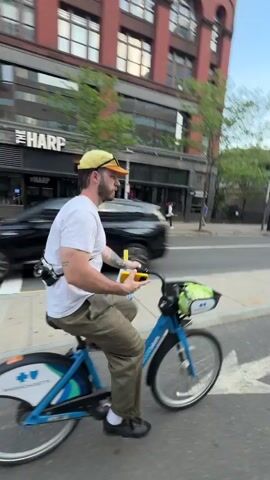 The video shows a person riding a blue bike on a city street. The rider is wearing a white t-shirt, khaki pants, and a yellow cap. There is a white bottle in the bike basket. The person rides past a tall red brick building with many windows. 

The camera's view shifts to show the rider ahead, biking in a designated bike lane.  The bike lane is marked with a yellow line.  Buildings are visible on both sides of the street.  The rider is now wearing a blue shirt and has an orange bag slung over his shoulder.  As he approaches an intersection, a "No Left Turn" sign is visible. A dark car waits at the red light.