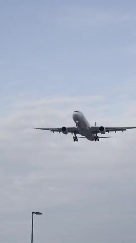 An airplane is flying through a cloudy sky. The airplane is a commercial airliner with two engines mounted under its wings. The landing gear is extended, suggesting it is either preparing to land or has just taken off. The plane is angled upwards, and its lights are on. The sky is overcast with white clouds, and there is a hint of blue sky peeking through. In the foreground, a lamppost is partially visible, indicating the shot was taken from the ground. The overall impression is one of a routine aviation event.