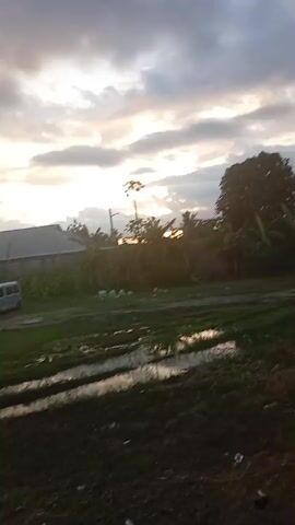 The video shows a rural landscape under a cloudy sky with the sun peeking through. In the foreground, there's green grass with puddles of water, and some litter, like white fabric or paper. A motorcycle partially covered with a white sheet is visible. In the background, a dirt road runs horizontally with several parked vehicles of various types, including cars, vans, and SUVs. Behind the vehicles is a long, light-colored building with a corrugated metal roof. Vegetation, including trees and bushes, lines the back. A water tower can be seen in the background to the left.  The overall atmosphere is somewhat gloomy due to the overcast sky.