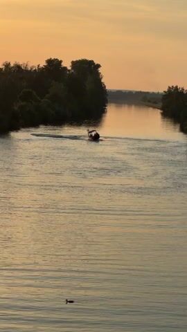 A boat is moving down a river during sunset. The boat is red and white and has people on it. The river is calm and the sky is orange and yellow. There are trees on the bank of the river.