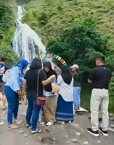 The video shows a group of people gathered at the base of a tall, cascading waterfall surrounded by lush green foliage. The scene appears to be a tourist attraction, with a colorful archway displaying text that is partially obscured but seems to read "AIR TERJUN". The people are dressed casually, suggesting a daytime visit. Some are looking at the waterfall, while others are interacting with each other. The overall mood seems relaxed and enjoyable, typical of a scenic outing. The presence of a waterfall and the dense greenery point towards a natural park or a rural area. The weather appears to be pleasant, with no indication of rain or extreme conditions. The location is identified as Sosor Dolok, Indonesia, which is known for its natural beauty, including waterfalls.