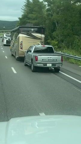 The video shows a silver Ford F-150 pickup truck towing a large RV on a highway. The RV is beige and brown with the "Tiffin" logo visible on the back. The truck and RV are in the right lane of a multi-lane highway. There are other vehicles in front of them, including another RV and a white passenger vehicle. The road is surrounded by trees and hills, suggesting a rural or scenic location. The sky is overcast. The camera appears to be mounted inside another vehicle, looking forward through the windshield. The video is very short, showing only a few seconds of footage.