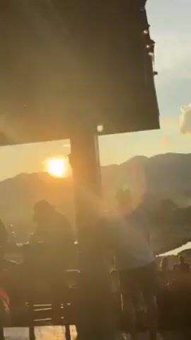 The scene is an outdoor gathering at sunset with a view of mountains and fields. A group of people is standing on a hilltop, watching the sunset over a scenic valley with fields and a small river. The foreground is partially shaded by a structure with a string of lights. The sun is shining brightly, causing lens flare. The sky is blue with some fluffy clouds. People are standing, looking at the view, some taking pictures. The atmosphere is relaxed and scenic.