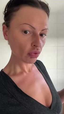 A woman is applying lip gloss, as a base, to her lips. She holds a silver tube of lip gloss in her right hand. She is wearing a dark gray v-neck shirt. Her hair is pulled back. The background is white tile. The video is a close-up shot.