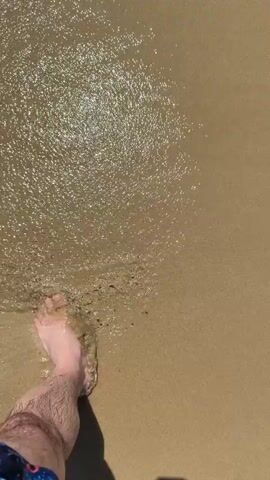 The video shows a first-person perspective of someone walking on a sandy beach. Their bare feet are visible, with the right foot stepping forward into shallow water. The water is clear and reflects the sunlight, creating shimmering patterns on the sand. The person is wearing patterned swim trunks. The overall scene is one of a relaxed walk along the shore.