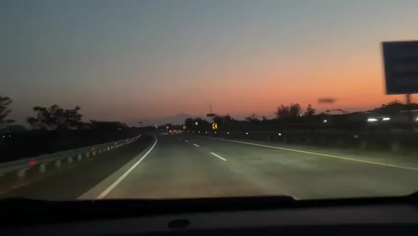 The video is a first-person view from inside a car driving on a highway at dusk. The sky is a gradient of deep blue to orange, with a distant mountain range silhouetted against the horizon. The highway is paved and well-lit with streetlights. There are guardrails on either side of the road, and a few other cars are visible in the distance. A black SUV is driving in the same direction as the camera car, slightly ahead and to the right. The SUV's brake lights are illuminated. The camera car's headlights are on and illuminating the road ahead. The overall impression is a smooth, steady drive on an open highway as the day transitions to night.