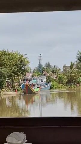 The video shows a wide shot of a river with boats docked on the bank. The water is murky brown. On the left bank of the river, there are trees and vegetation, with a small wooden structure. Two boats are visible, one larger blue and white fishing vessel and a smaller, darker boat with a yellow trim. The larger boat is docked alongside the smaller boat. In the background, there are buildings and a tall tower structure. The sky is overcast. The video is framed by a dark border at the top and bottom, suggesting it was filmed through a window or opening.
