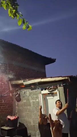 The video shows two men and a child in what appears to be a backyard at dusk. The child is on the left, holding a sparkler. The man on the right is holding a sparkler and looking up at the sky. The man in the middle is also holding a sparkler and looking up. There is a house in the background with a red-tiled roof. There are also some trees with green leaves at the top of the frame. Smoke is visible in the air, indicating that fireworks are being set off. The sky is a deep blue, suggesting that it is nighttime or late evening. The overall scene depicts a festive or celebratory occasion.