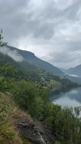 The video pans across a misty, mountainous landscape. On the left, a steep, green mountainside is dotted with small houses and farms. The mountains are covered in lush vegetation, and clouds cling to the higher peaks. To the right, a large body of water, likely a fjord, reflects the overcast sky. Trees and rocky terrain are visible in the foreground. The overall atmosphere is serene and somewhat dramatic due to the weather conditions.