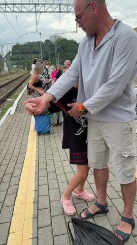 A man in a light gray shirt and beige shorts stands on a train platform, embracing a young girl. The girl has long, braided hair with red, green, and white strands. She wears a black t-shirt with a white graphic and a red and black plaid skirt, along with pink Crocs. The man has on dark sunglasses and dark sandals. Behind them, other people and luggage are visible on the platform, with train tracks and overhead wires in the background. An umbrella is on the ground at the man's feet.