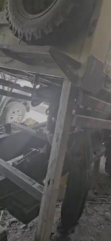 A video shows a person in dark overalls working under a damaged vehicle. The vehicle appears to be upside down and is supported by wooden beams. In the background, a white SUV is visible. The scene is dusty and appears to be outdoors.