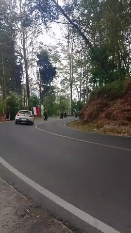 The video shows a curved asphalt road lined with tall trees and vegetation. Several vehicles are present. In the foreground and mid-ground, a black pickup truck drives along the road. To the left, a white car is also visible. A person on a motorcycle or scooter, wearing a blue jacket and a red and white flag attached to the back of the vehicle, is seen riding past the pickup truck and continuing down the road. Further in the distance, another motorcycle is visible. The sky appears overcast. The scene is shot from a low angle, possibly from a moving vehicle.
