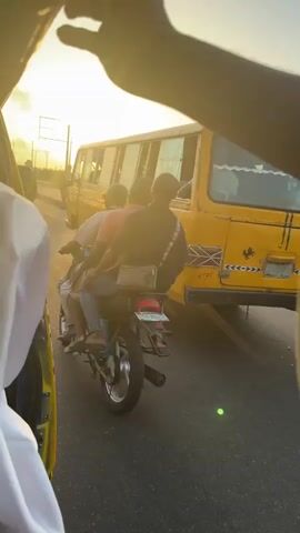 A low-angle, medium shot filmed from inside a vehicle shows traffic in what appears to be Nigeria.  The scene is bathed in the bright light of the setting sun.  A yellow bus with the word "LASHGHKY" painted on its side is in the right of the frame, along with other cars and buses in the distance. To the left, a yellow vehicle similar to the one the camera is in is partially visible.  In the foreground, a motorcycle carrying two people is stopped in traffic.  Their faces are not visible. The motorcycle has a license plate that reads "MTM 721 TE."  Two hands reach up and partially obscure the view.  The hands are dark-skinned and appear to belong to the person filming.  The overall atmosphere is one of a busy street scene during sunset.