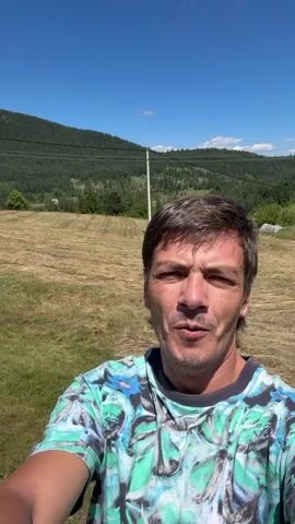 A man in a tie-dye shirt stands in a grassy field under a bright blue sky. He holds up his left hand, displaying a peace sign with his index and middle fingers. Behind him, a landscape unfolds with rolling hills covered in trees and dry grass, a few scattered houses, and a power pole. The man looks directly at the camera, with an open mouth as if speaking or exclaiming. The video appears to be a selfie, with the camera moving slightly to capture different parts of the background.