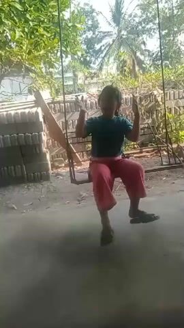 A child sits on a swing, wearing a teal t-shirt and pink pants. The child is in an outdoor setting, with trees and vegetation visible in the background. The swing appears to be homemade, with a wooden seat suspended by ropes from an unseen support. The child is moving the swing back and forth with their legs. The ground around the swing is dirt and there are piles of bricks to the left of the frame. The lighting suggests it is daytime.