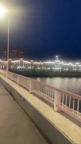 The video begins with a view of a bridge railing, partially obscuring a body of water. Beyond the water, a long building complex is illuminated by numerous lights, reflecting on the surface of the water. The sky is dark blue, indicating it is either dusk or nighttime, with a small, reddish-orange circular object, possibly the moon or a distant light, visible in the sky above the building. A small boat with a dark canopy is present on the water. The building complex appears to have a repetitive architectural style with multiple windows and decorative lighting. The overall scene suggests an urban or riverside environment at night.