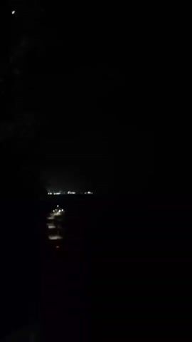 The media file captures a dimly lit scene at night, likely in El Tigrillo, Venezuela. The foreground is dominated by darkness, with a silhouette of foliage on the left side and a hint of a railing or structure on the bottom right. In the middle ground, a road or pathway is visible, illuminated by the headlights of passing vehicles, which appear as streaks of light due to motion blur. Further in the distance, a faint line of lights suggests a town or settlement. The overall ambiance is quiet and serene, with the darkness emphasizing the artificial lights that punctuate the landscape. The time of day is clearly night, and there are no discernible weather conditions beyond darkness. The scene evokes a sense of solitude and the quiet beauty of a rural or semi-rural area at night. There is no visible text in the media file.