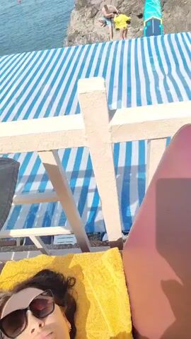This video is a selfie taken by a woman lying on a lounge chair outdoors. She is wearing a green and white polka dot bikini and dark sunglasses. She is resting on a yellow towel, and the lounge chair appears to be on a deck or patio. In the background, there is a table with some items on it, including a plastic bag with food and a pair of yellow flip-flops. Further in the background, there is a white building and some greenery. The lighting suggests it is daytime, likely sunny. The woman is looking at the camera and appears relaxed and enjoying the outdoors. There is no visible text in the video.