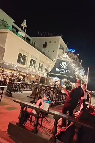 The video captures a nighttime outdoor scene at a venue called Bounty Rimini, decorated with a pirate theme. A band is performing on a raised wooden platform that resembles a ship's deck. A guitarist wearing a hat and a black shirt stands center stage playing an electric guitar. To his right, a drummer in a green shirt plays a drum kit. A third musician, possibly a keyboardist, is partially visible behind the drummer. In the foreground, a figure in a pirate costume sits on a stool, holding a sign that reads "Bounty." The background features diners seated at tables under awnings, and string lights illuminate the area. The overall atmosphere appears lively and festive.
