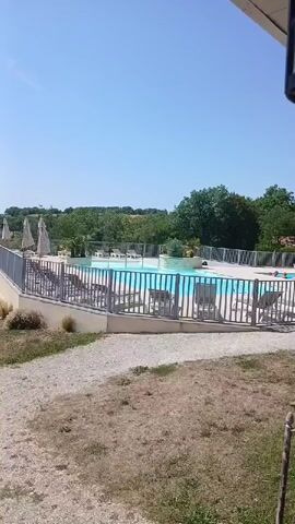 The video shows a drone shot flying over a swimming pool area on a sunny day. The pool is rectangular and has blue water. Several lounge chairs are around the pool, and some people are visible in the water. A fence surrounds the pool area, and beyond that, there are trees and rolling hills in the background. The drone then pans to the left, revealing more of the landscape.