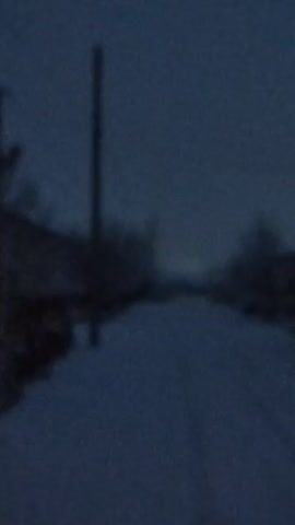 The video shows a blurred view of a snowy street at night. There are a few buildings with lit windows visible. The camera moves quickly, making it difficult to see details. There are some trees and a utility pole visible as well. The scene is dark and appears rural.